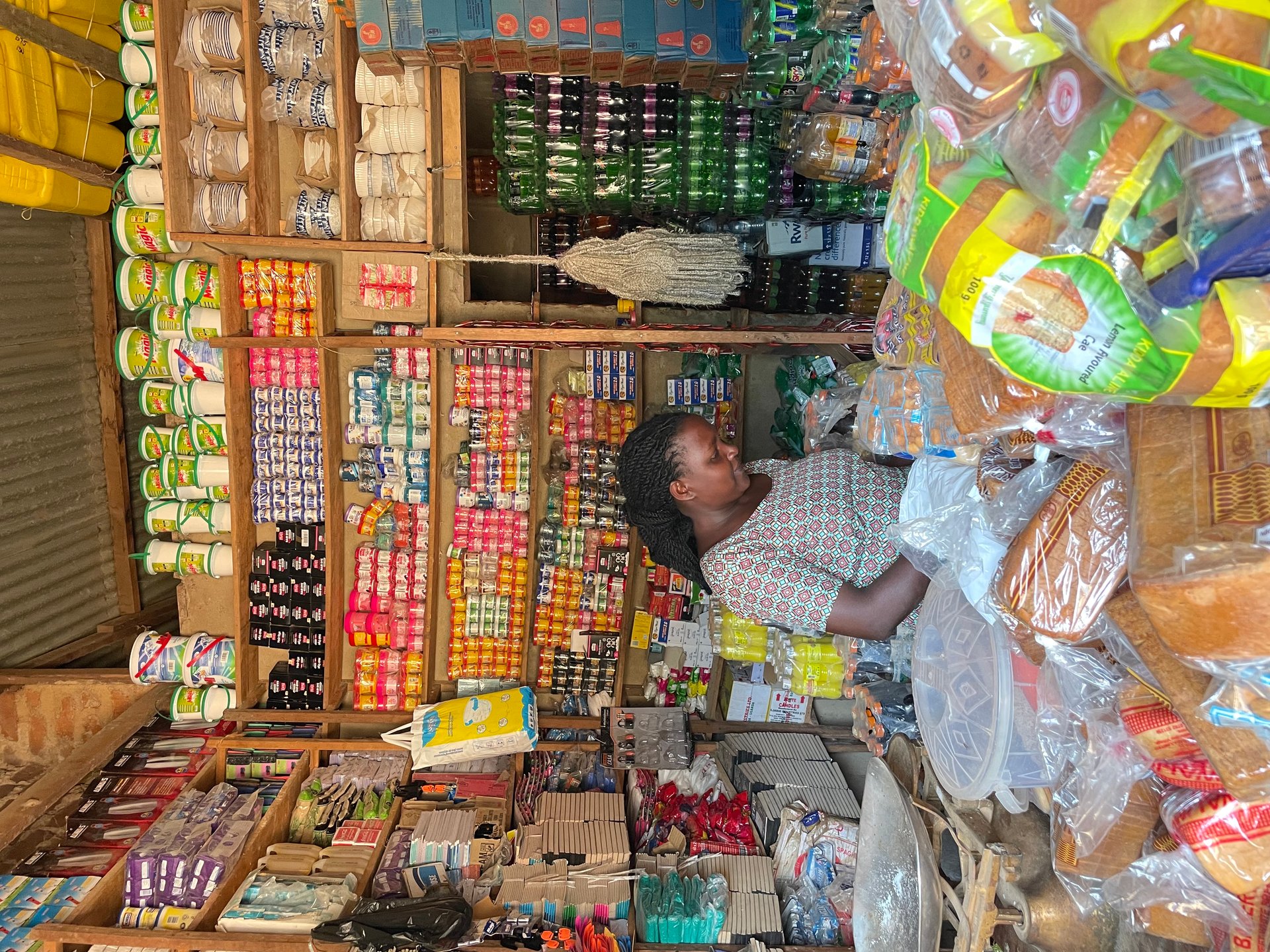 Local shopkeeper standing behind the counter of a densely stocked village store in Buhoma, shelves filled floor to ceiling