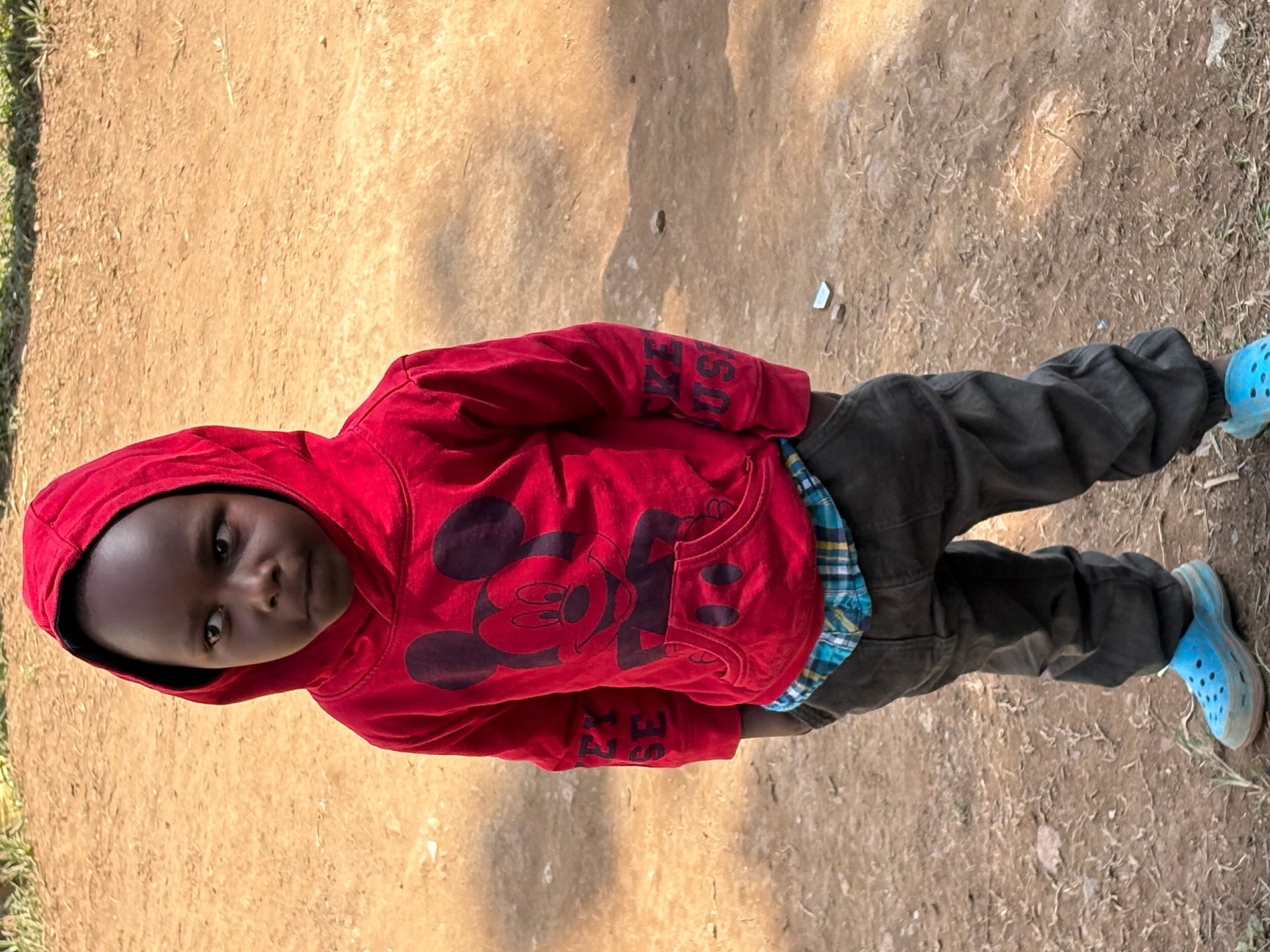 Young boy standing on red earth in Buhoma wearing a red Mickey Mouse hoodie, looking directly at the camera