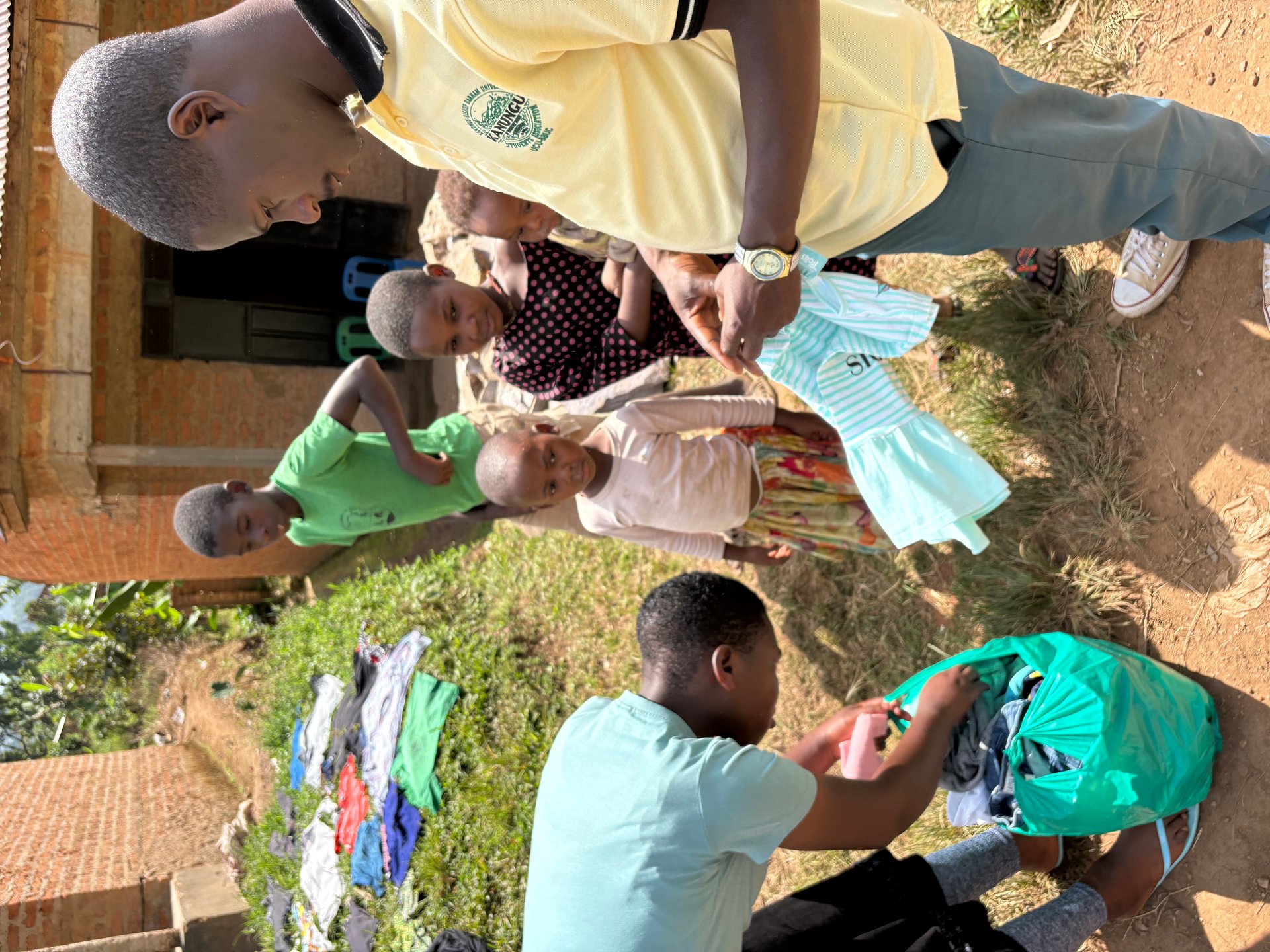Two young men sorting donated clothing from a green bag while children watch at the Buhoma orphanage