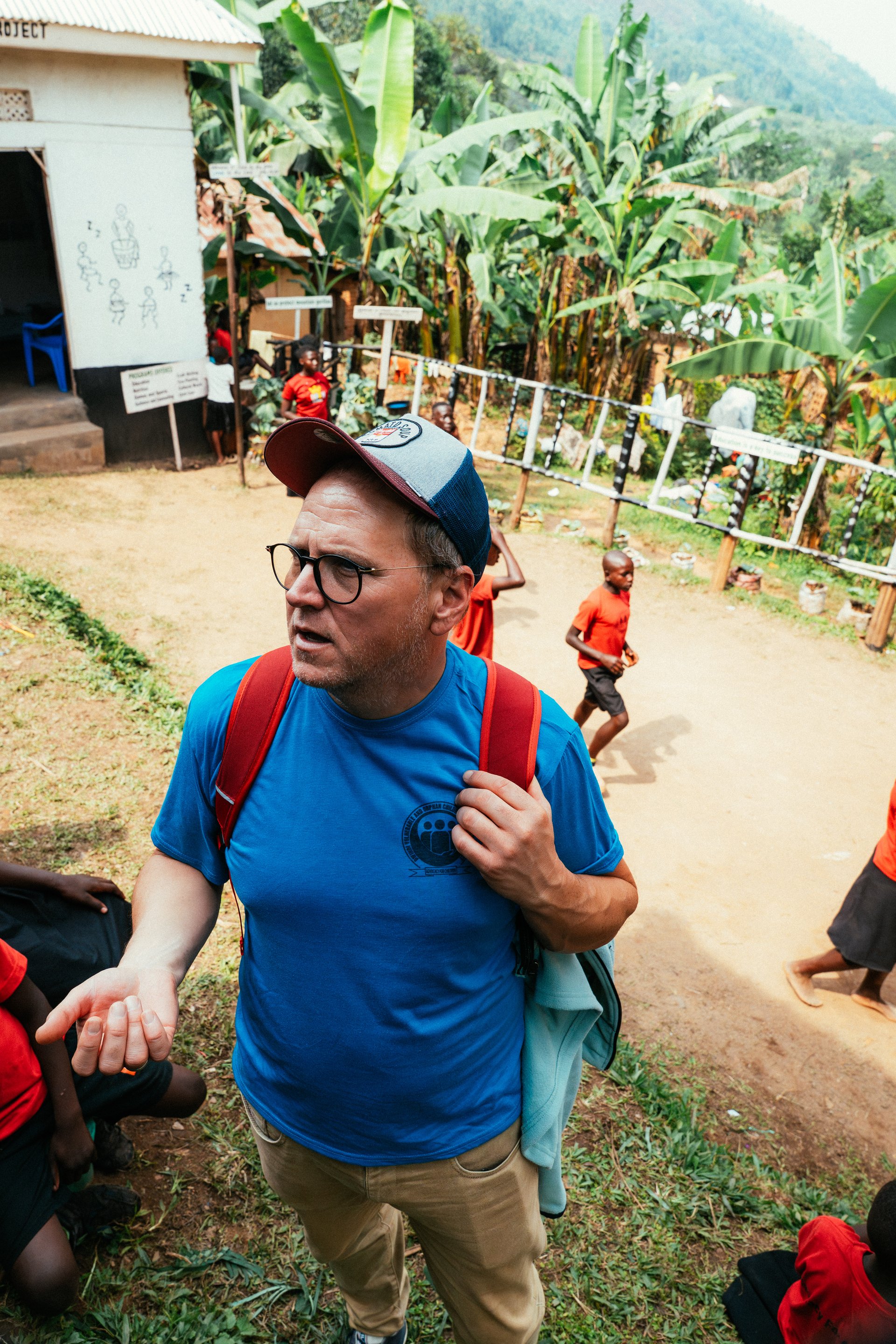 Mark Suer at a community project in Buhoma, surrounded by children in red school uniforms with banana plantations and green mountains behind