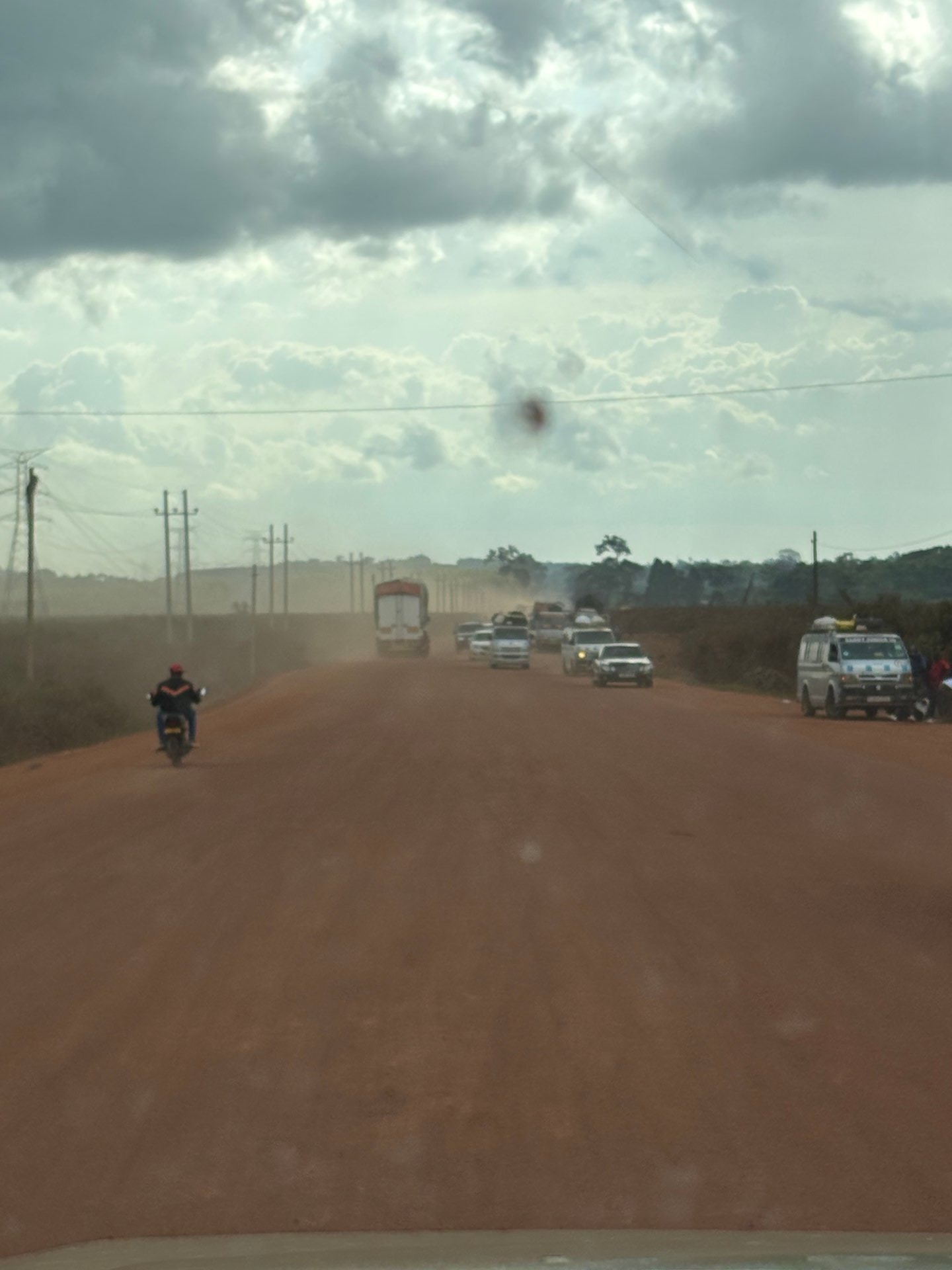 Red laterite road stretching into the distance with a BodaBoda rider, lorries, and power lines under dramatic clouds