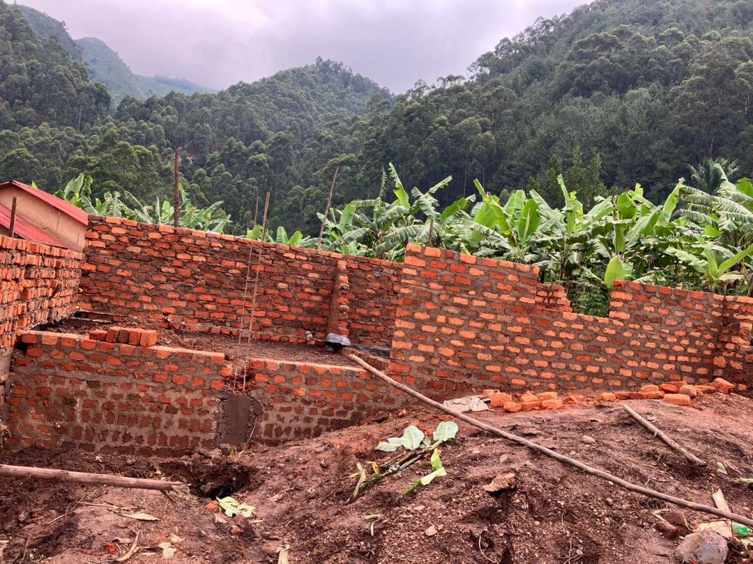 HopeKitchen foundation walls rising from red earth — hand-made red bricks with banana trees and forested mountains behind