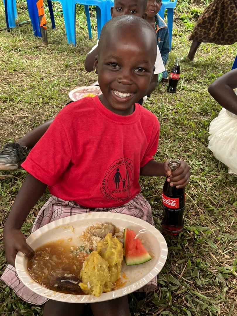 Smiling child in a red shirt holding a plate of food at a community event in Buhoma