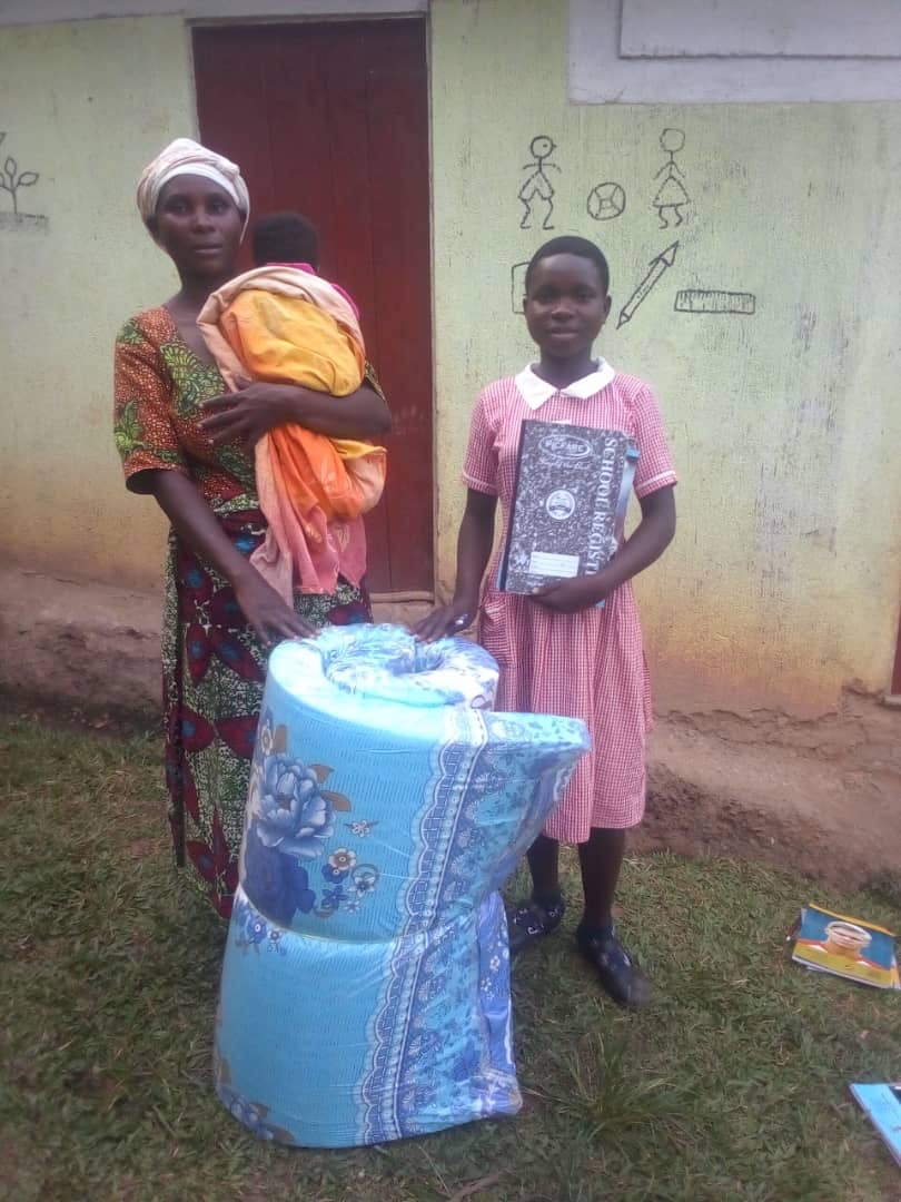 Mother with baby and schoolgirl holding a school register next to a new mattress in Buhoma