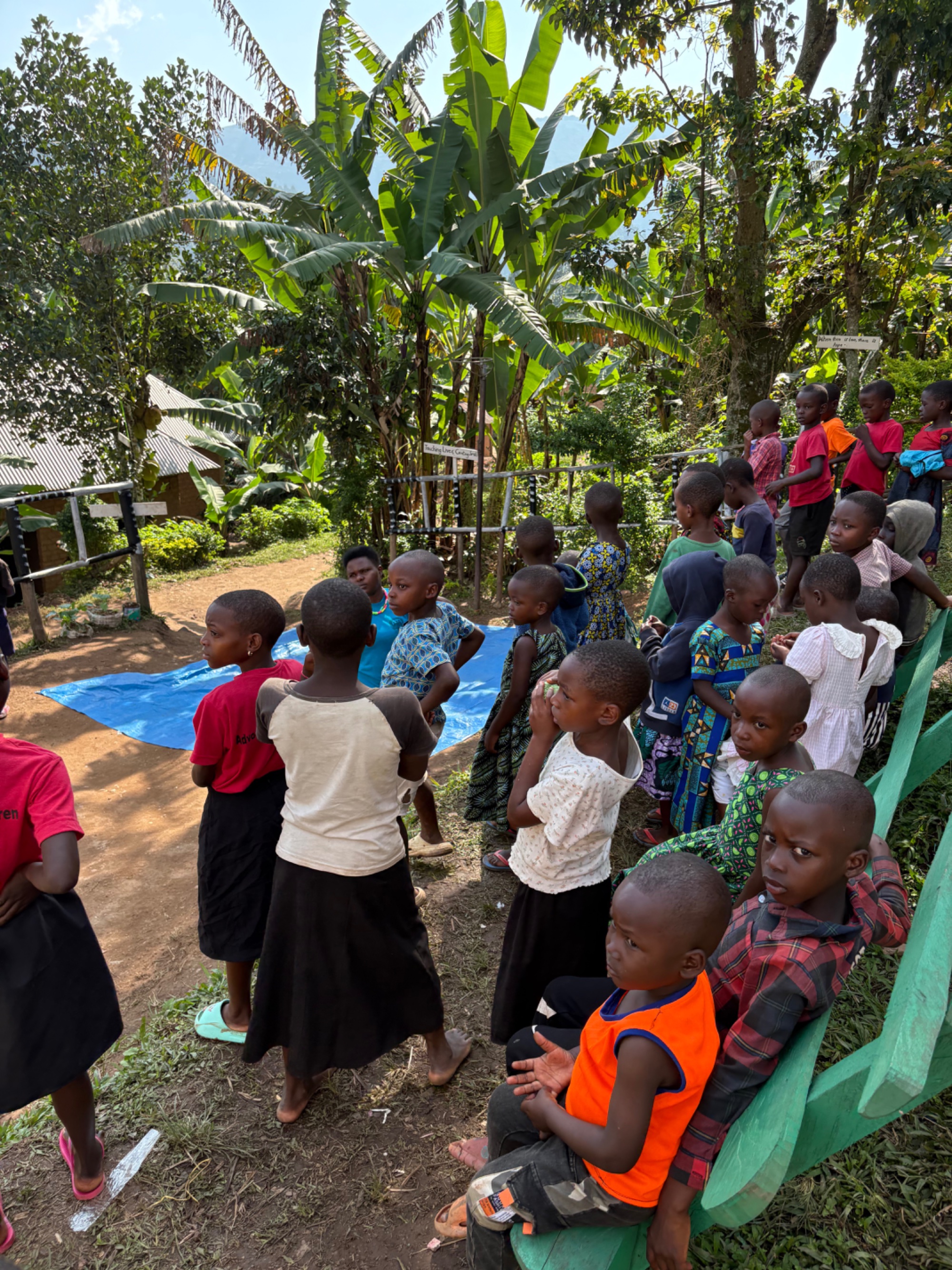 Dozens of children gathering under banana trees at the Buhoma orphanage, a blue tarpaulin on the ground