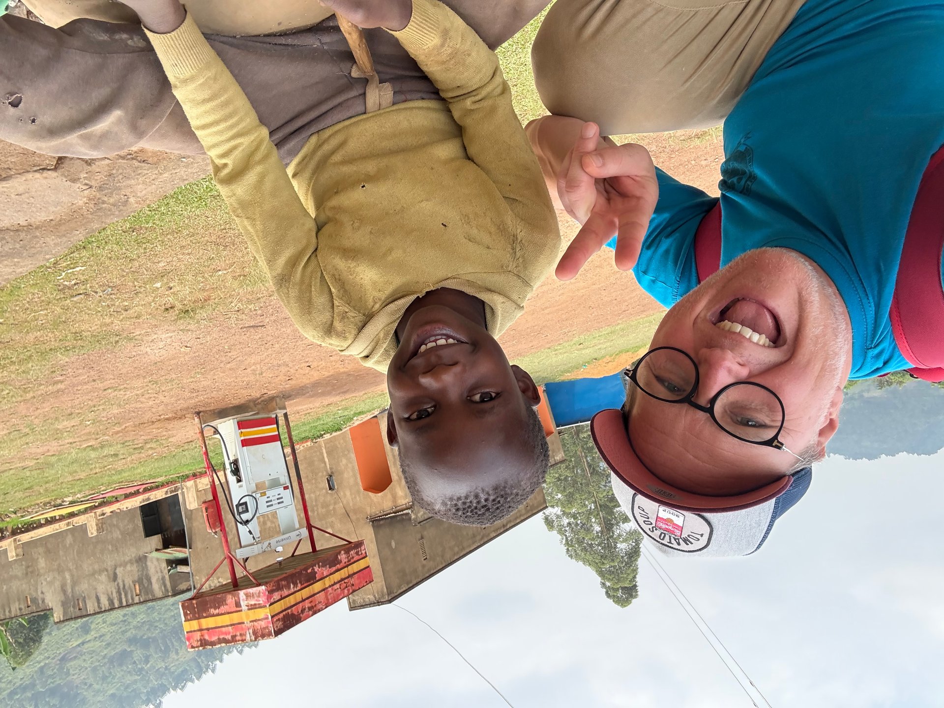 Mark Suer smiling with a local young man in front of a roadside kiosk on red earth in Buhoma